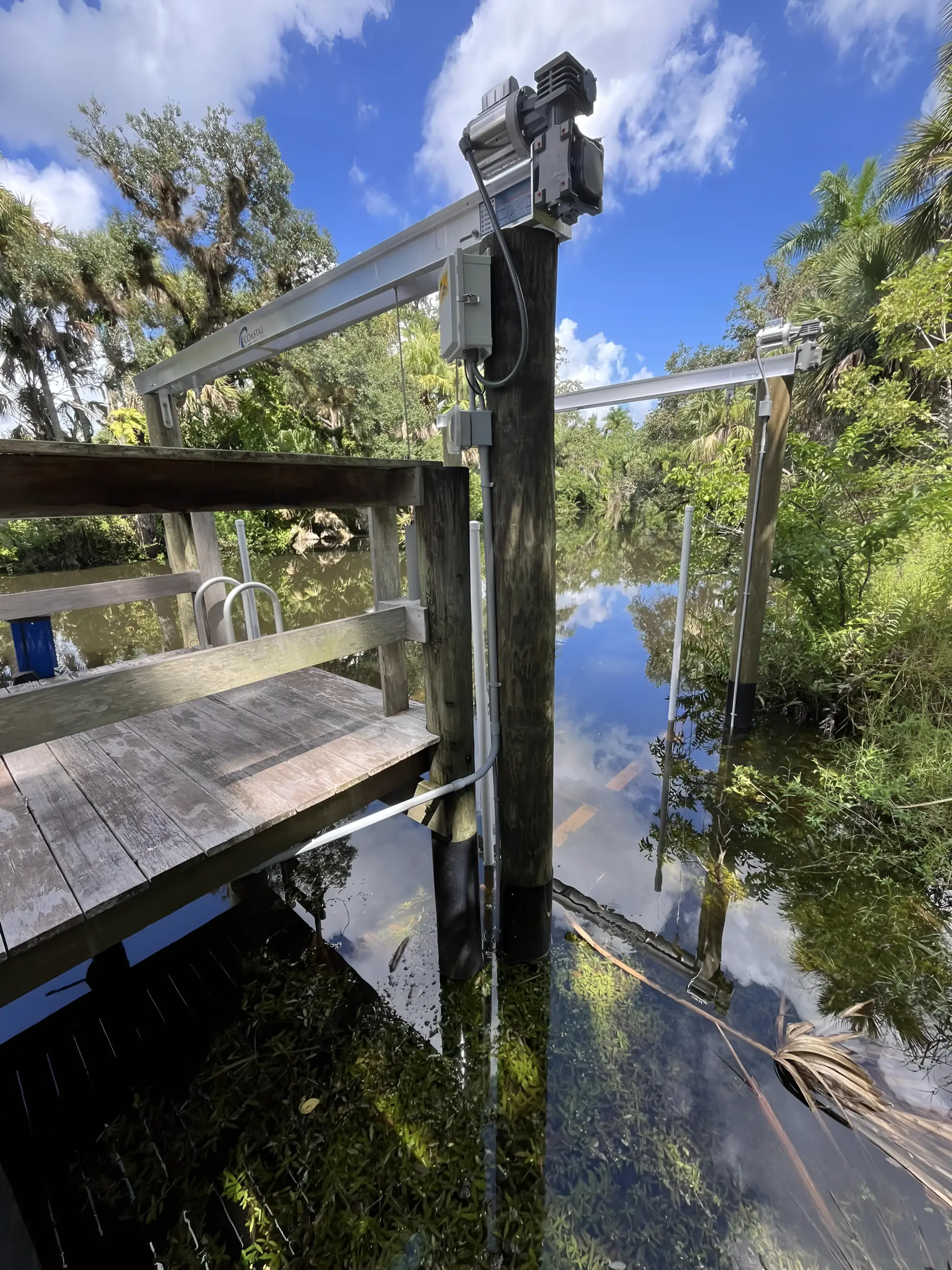 A wooden dock and aluminium boat lift on a mirror-flat creek under a blue sky, lift motors, control box and conduit all in view.