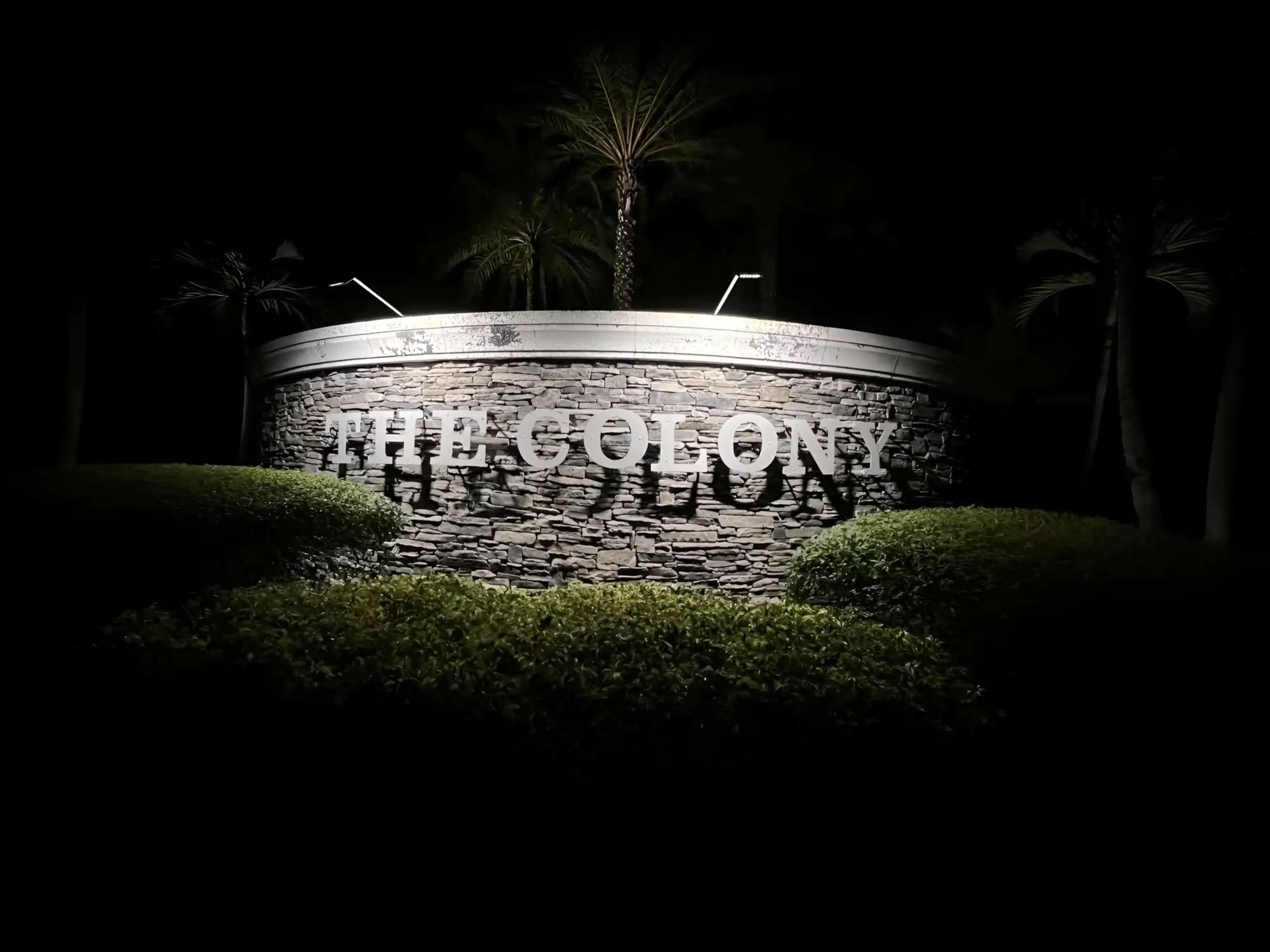 A stacked-stone community entrance monument washed by new landscape uplighting at night, with palms silhouetted behind it.