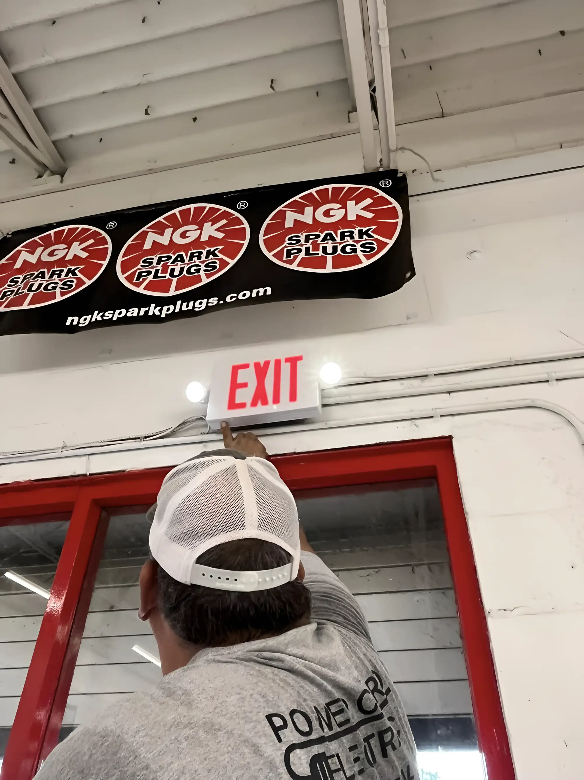An electrician in a Power Cords Electric shirt, seen from behind, pointing up at a newly installed combination exit sign and emergency light above a red shop door.