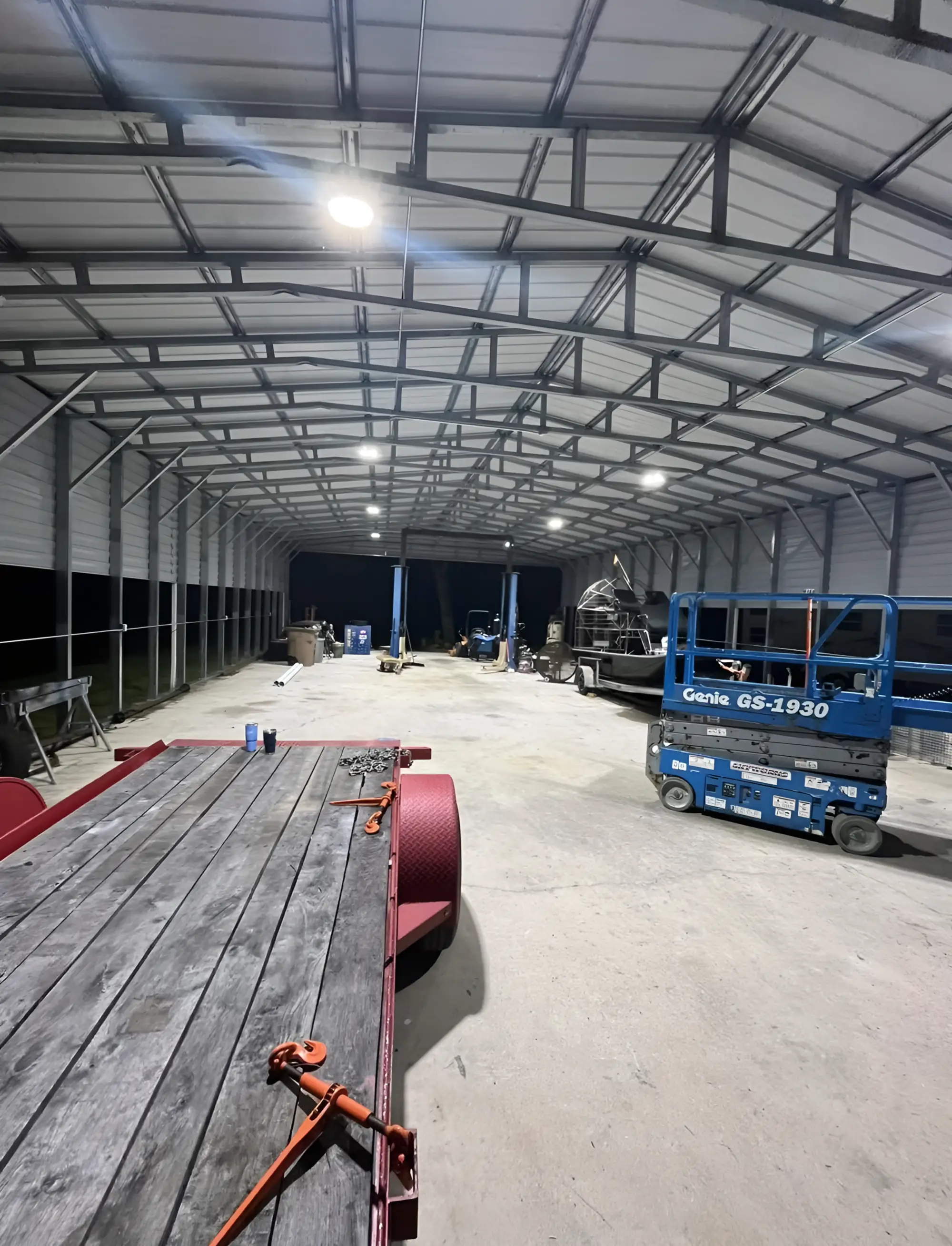 The inside of a large steel pole barn at night with new LED high bays lit across the truss ceiling, an airboat, a scissor lift and an equipment trailer on the slab.