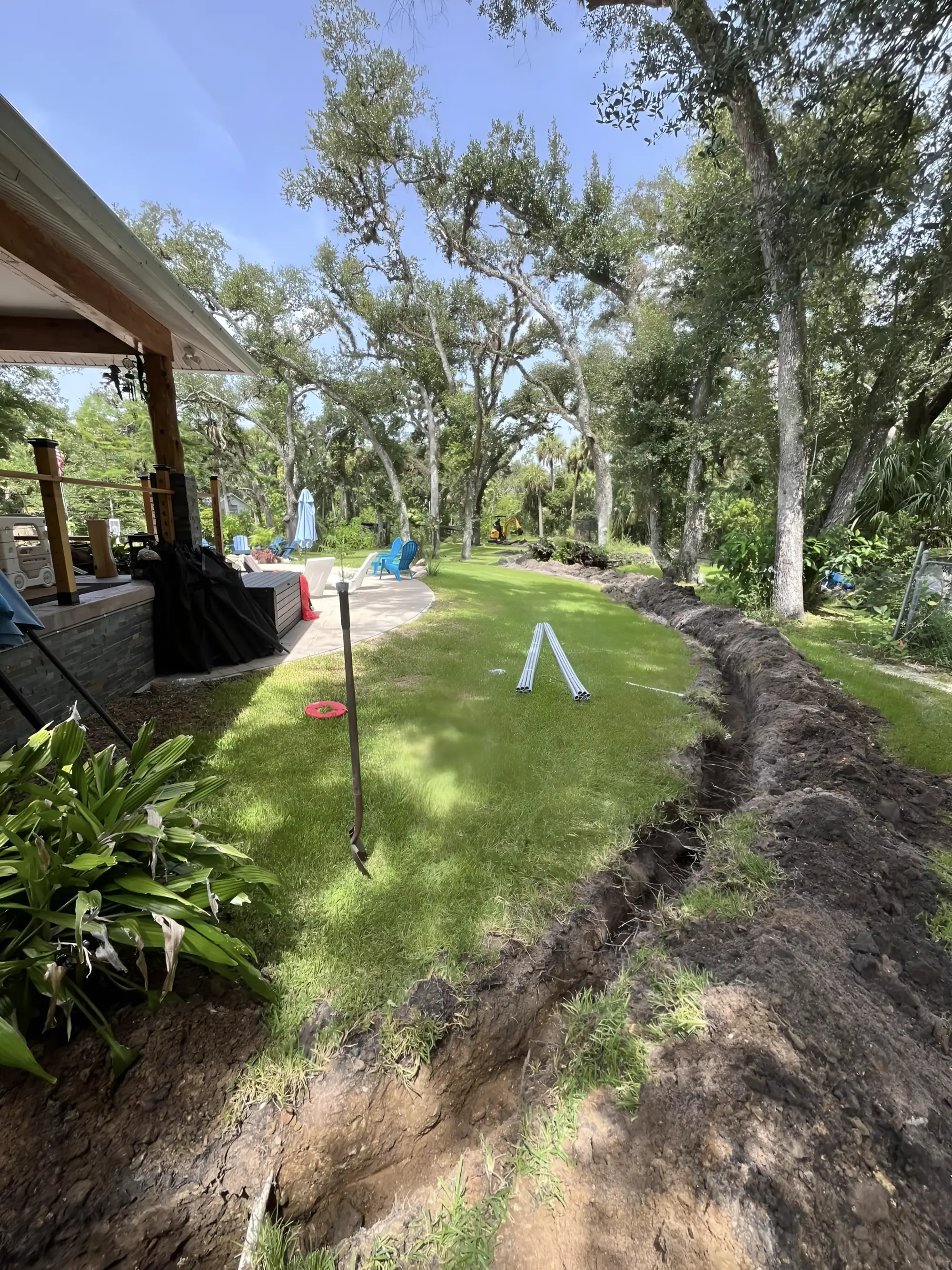 A freshly cut trench curving across a sunny backyard toward a paver pool patio, with bundles of grey PVC conduit staged on the lawn and a mini excavator among live oaks beyond.